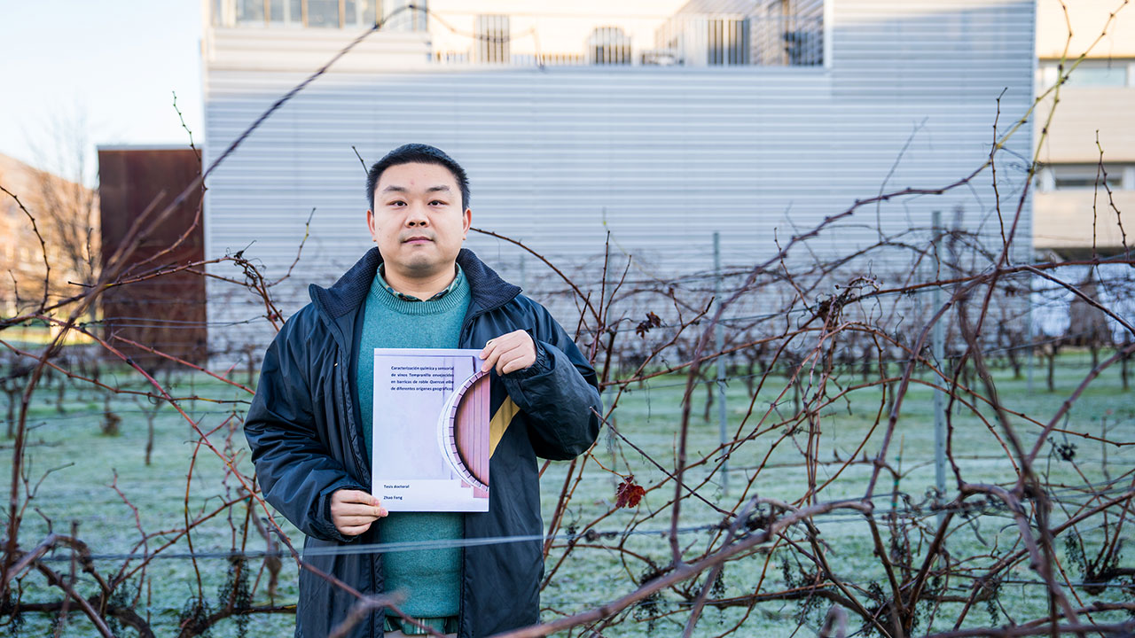 Zhao Feng posa con el volumen de su tesis en los viñedos y junto a la bodega experimental de la Universidad de La Rioja